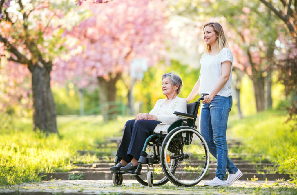 A caregiver is helping an elderly woman with dementia in a wheelchair to walk outside.