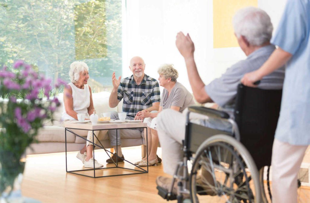 A group of happy seniors engaged in conversation together with their caregiver in a home setting.