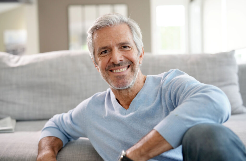 A senior man sitting on a couch, smiling and looking directly at the camera.