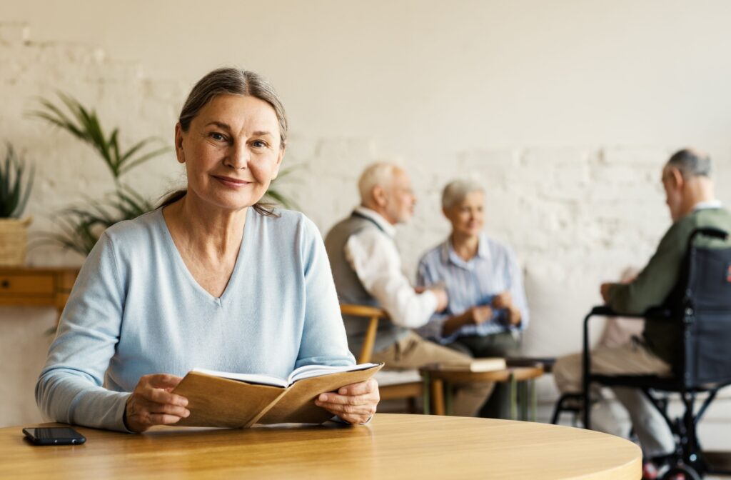 A senior woman smiling while sitting at a table holding a book with other seniors in the background