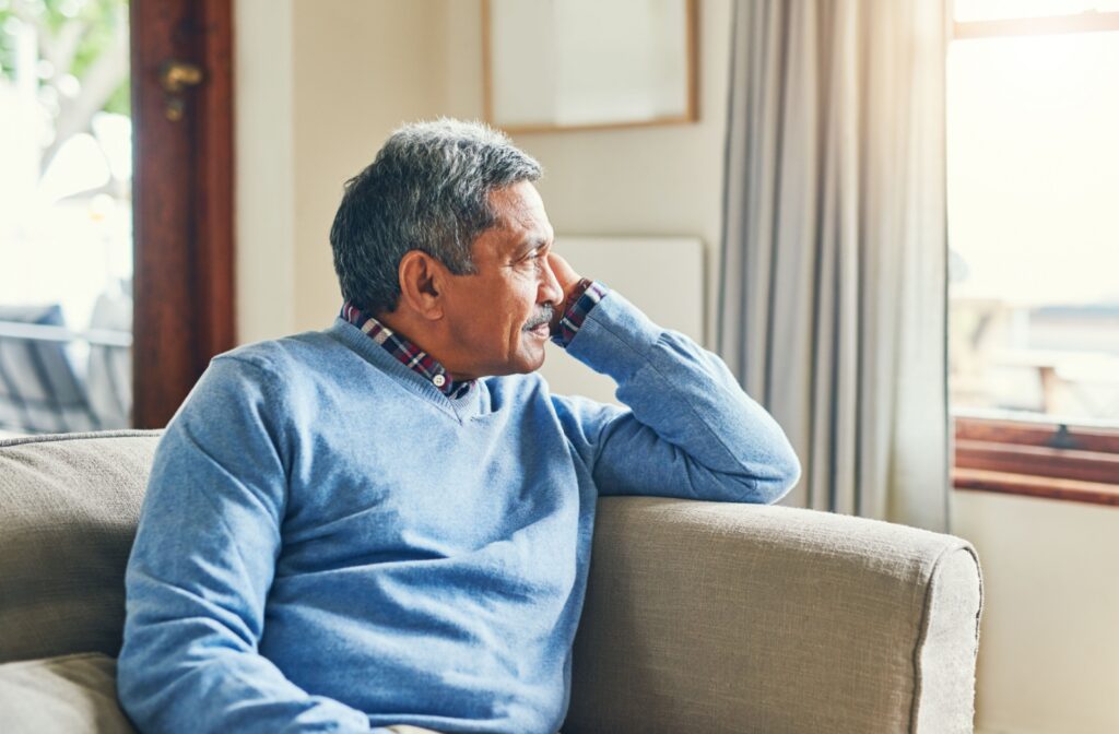 An older man in a blue sweater sits on a couch, looking out of the window.