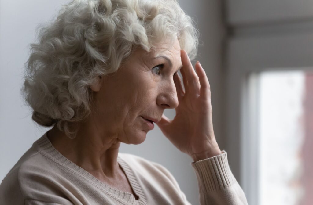 A worried older adult touches the left side of their face in confusion while standing in their living room